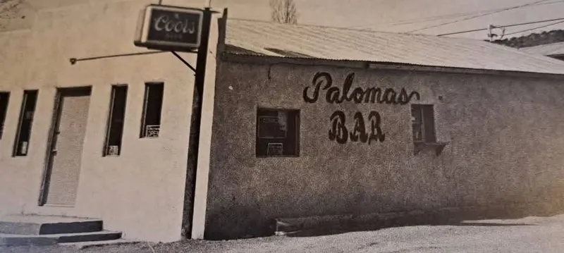 Old black and white photo of the Palomas Bar building: a stucco storefront with a Coors sign on a pole and Palomas BAR hand painted in script on the wall